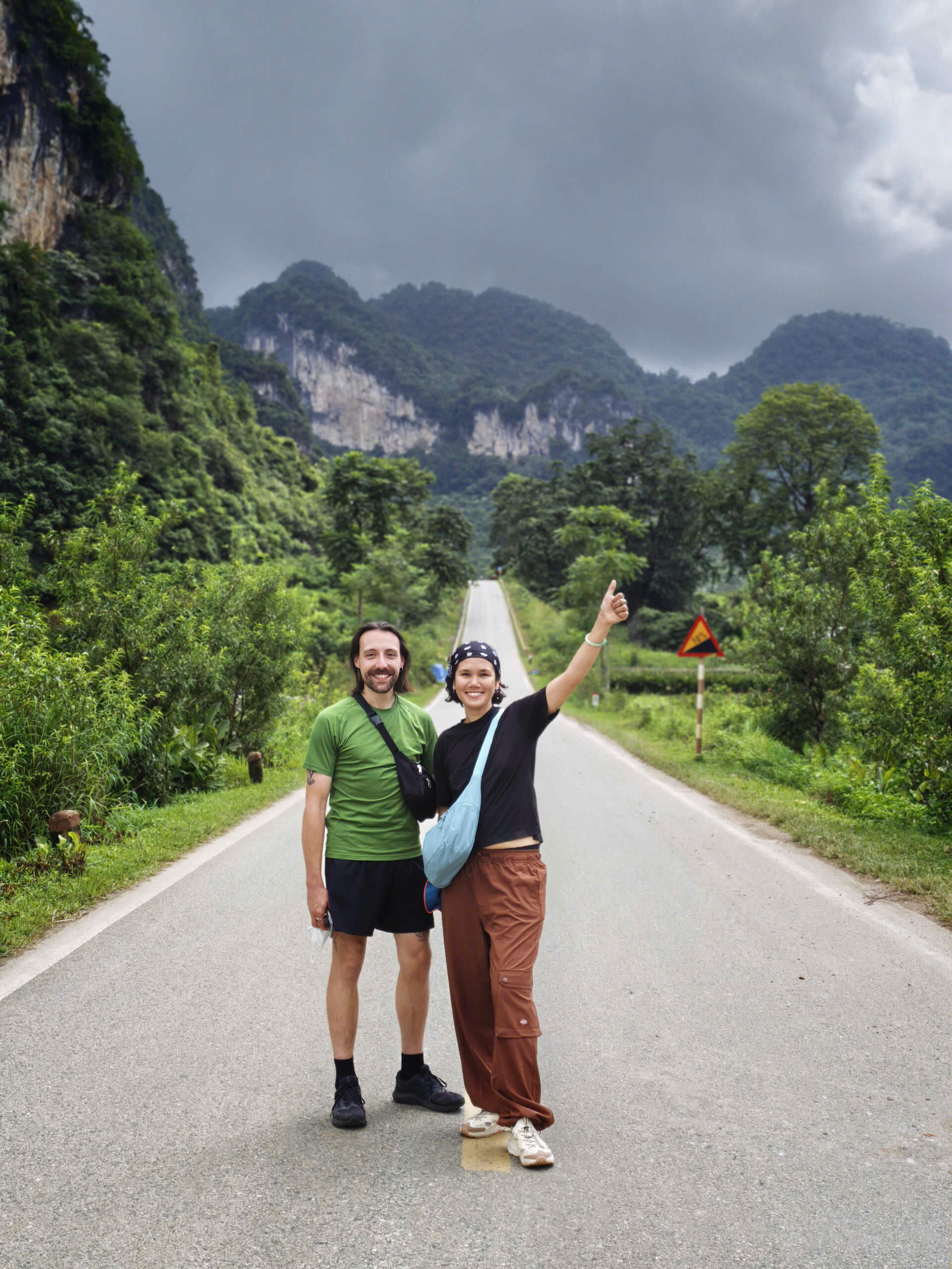 A couple posing happily in the middle of a scenic, empty paved mountain road surrounded by lush green valleys and towering limestone karst mountains on the Cao Bang Loop.