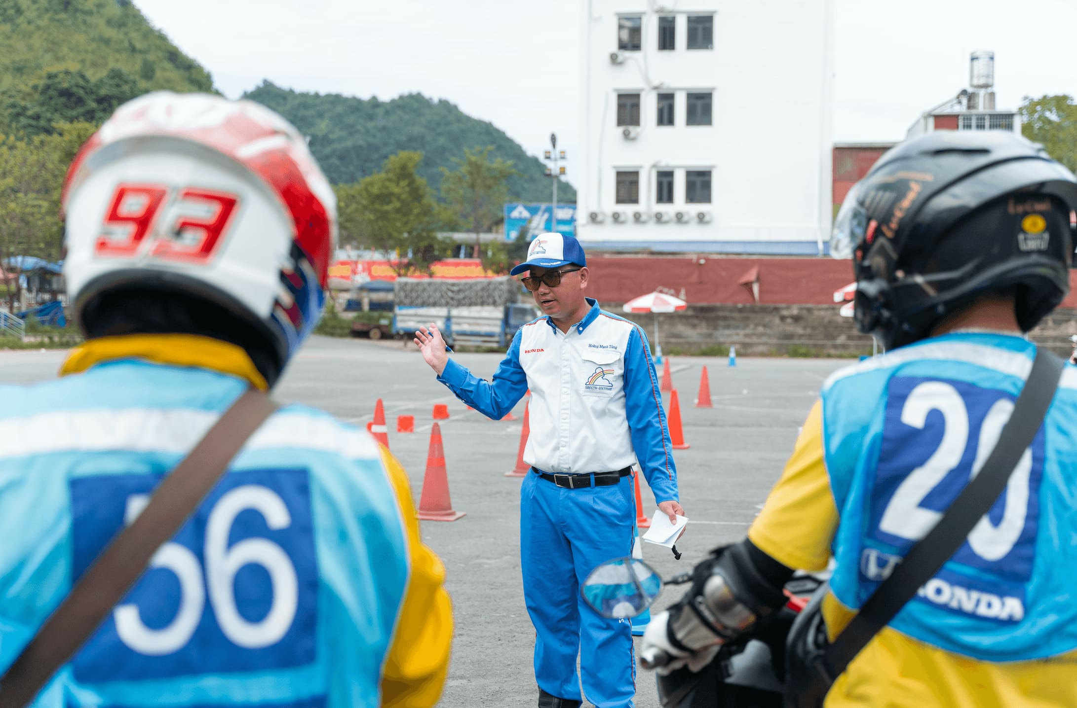 A Honda Vietnam safety instructor in a blue and white uniform gives direct, hands-on coaching to two Motorventures Easy Riders wearing helmets and yellow uniforms on a closed training course.