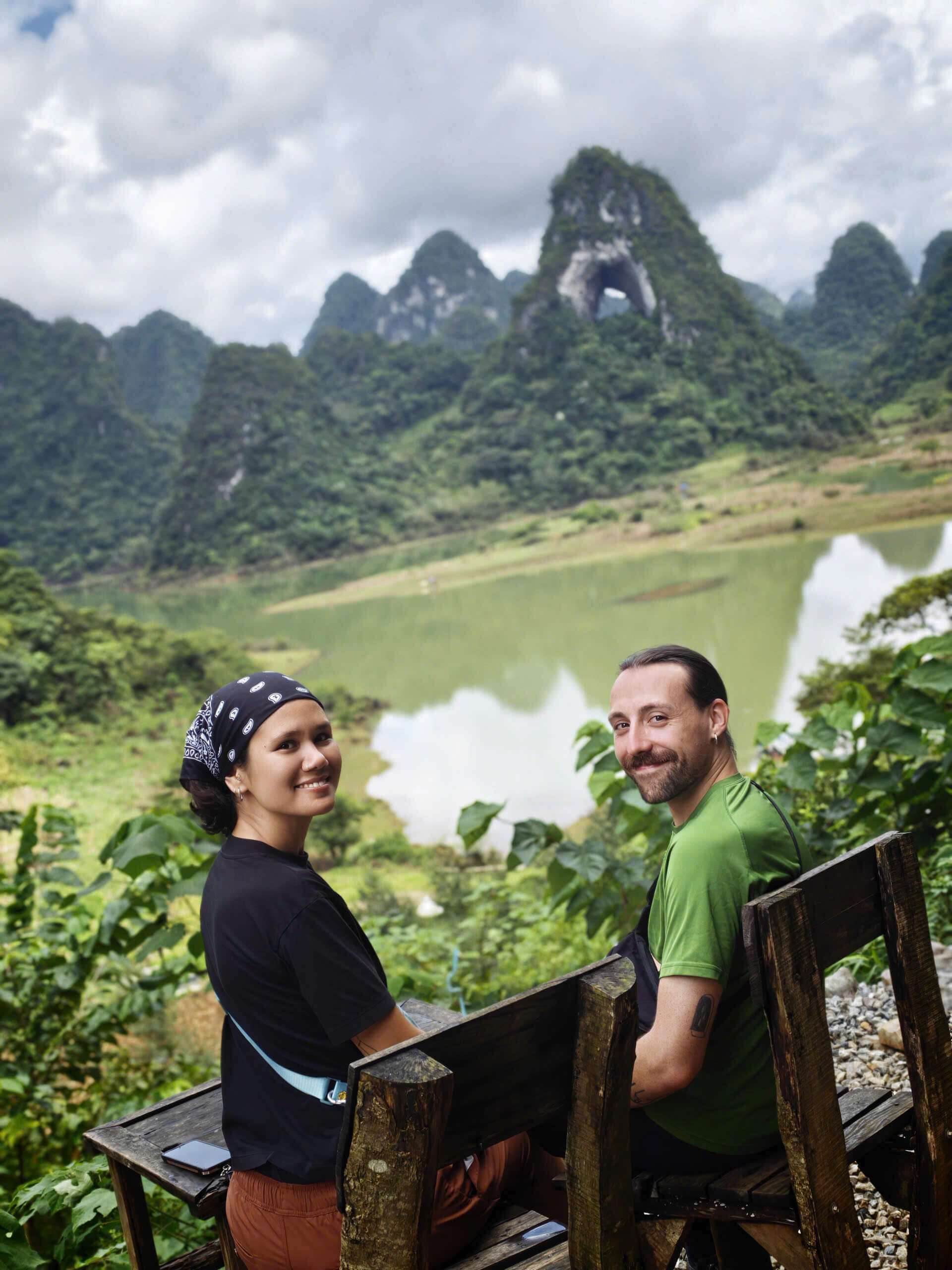A smiling couple sitting together on a wooden bench overlooking a calm green lake and the famous Angel Eye Mountain, a massive karst peak with a natural hole through it in Cao Bang.