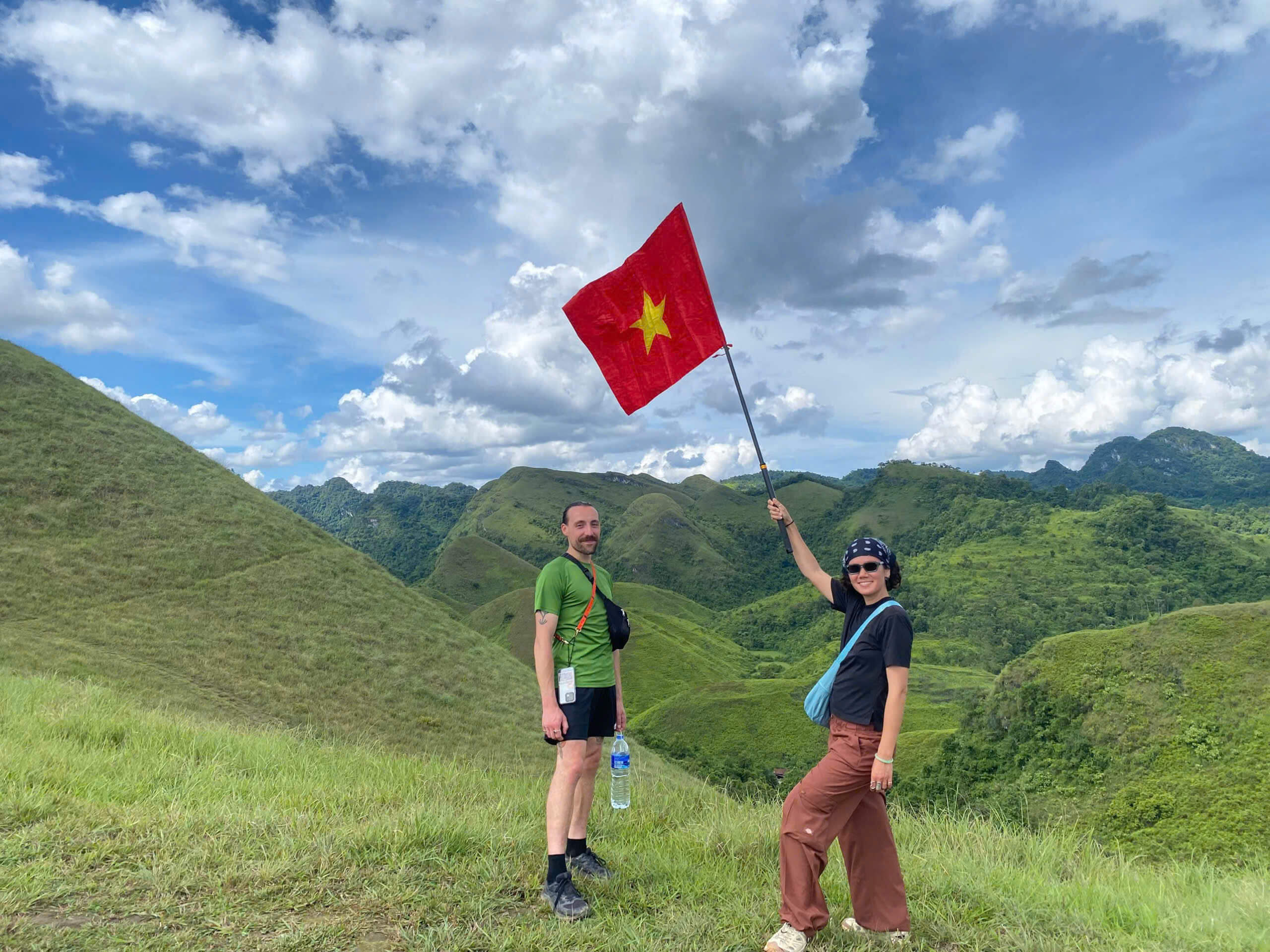 A couple standing on a lush, rolling green hill on the Cao Bang Loop, with the woman proudly waving a large red Vietnamese flag against a backdrop of endless green mountains and a blue sky