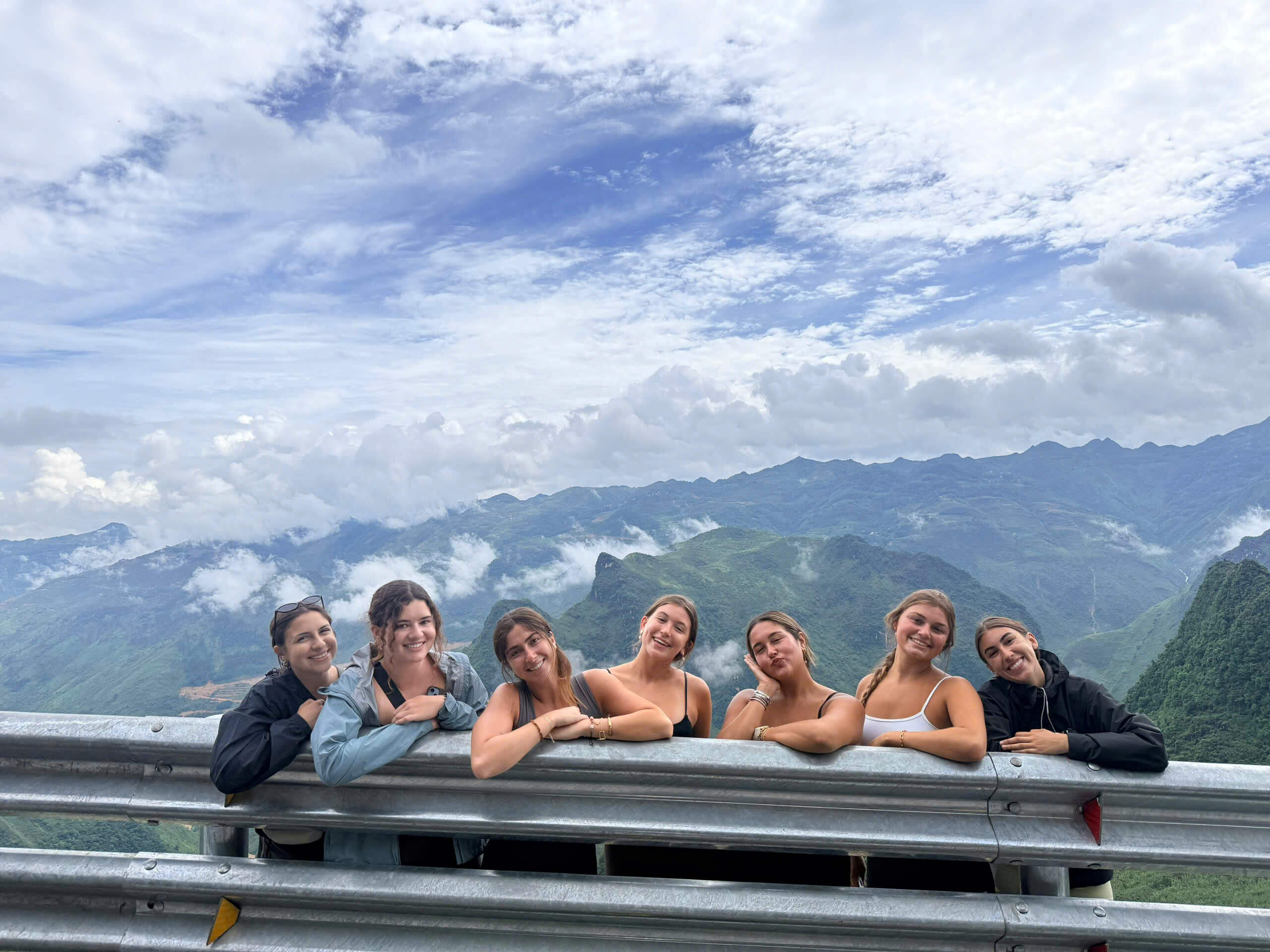 A group of seven female tourists leaning on a highway guardrail, smiling against a stunning backdrop of steep green limestone mountains and low-hanging white clouds in Ha Giang.