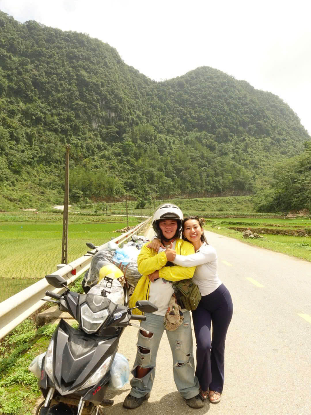 A happy female tourist hugging her Motorventures Easy Rider guide next to a motorbike on a quiet, perfectly paved road surrounded by green rice paddies on the Cao Bang Loop.