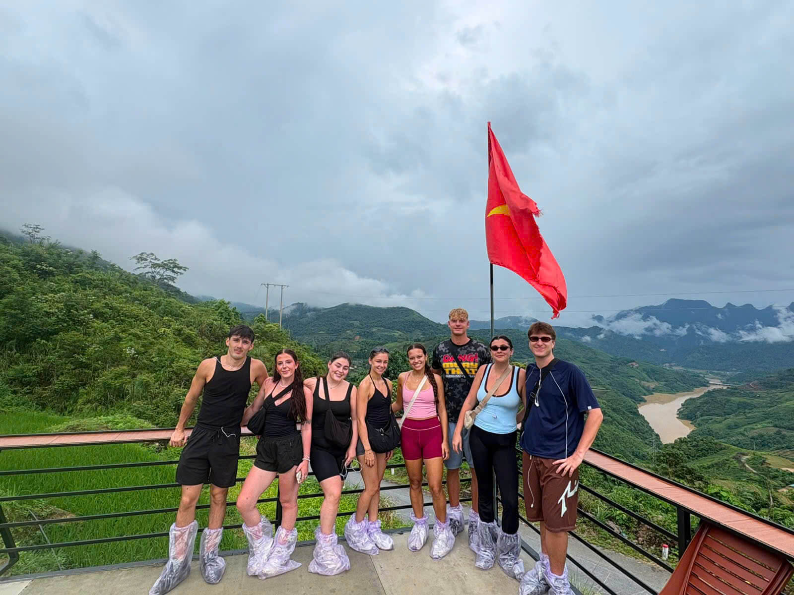 A group of tourists in protective rain shoe covers posing on a scenic observation deck overlooking a deep green valley and a winding brown river in Ha Giang, with a large Vietnamese flag flying.