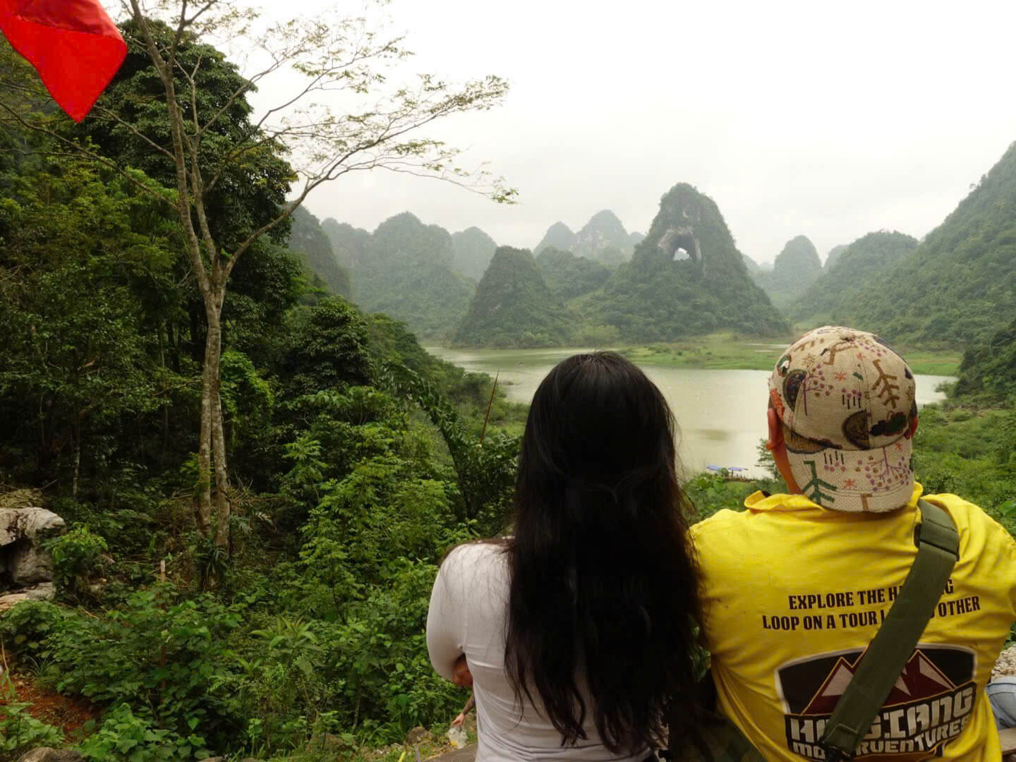 A female tourist and her Motorventures guide in a yellow shirt looking out over a lush green valley and lake towards Angel Eye Mountain in Cao Bang.