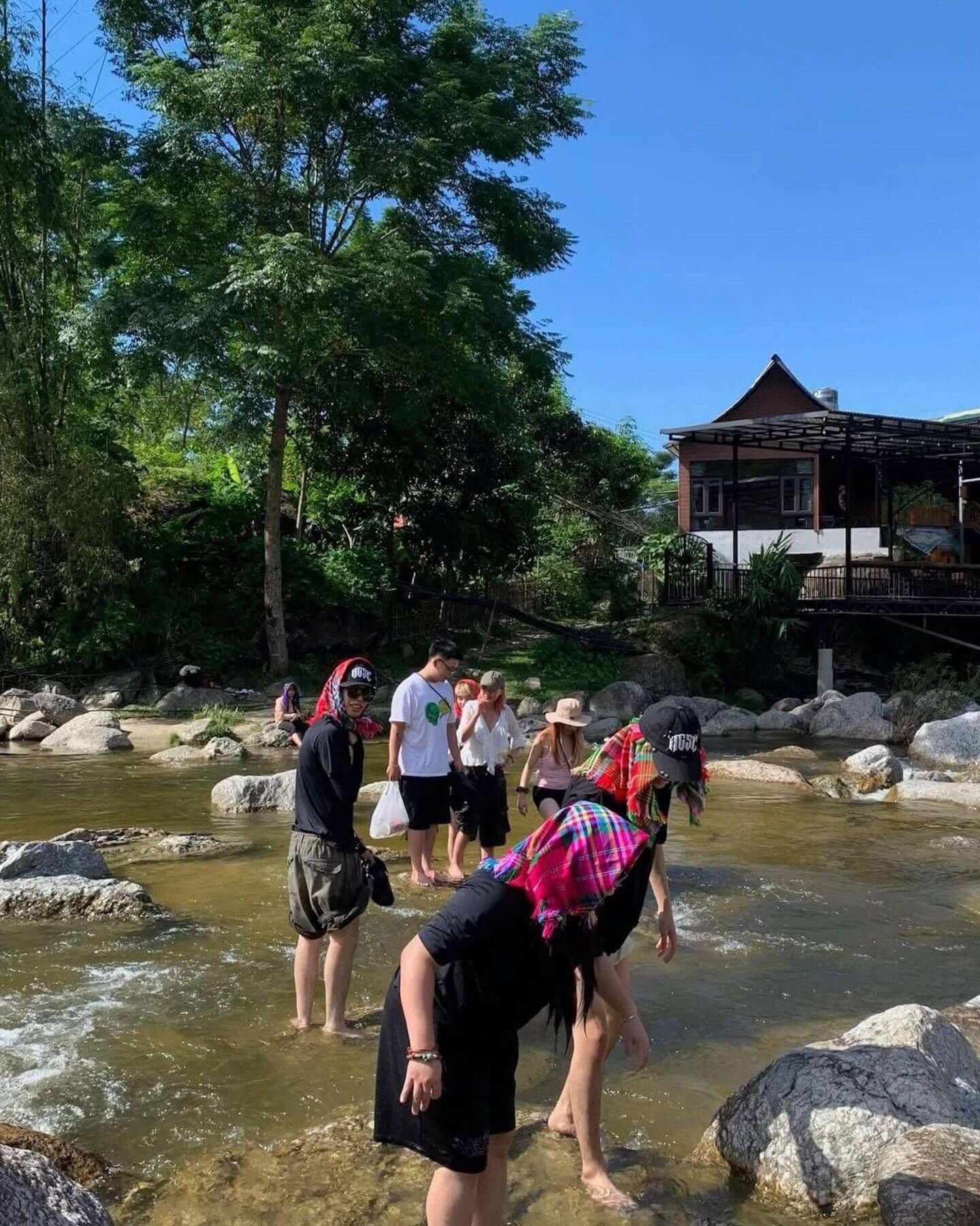 A group of tourists, some wearing traditional colorful ethnic headscarves, playfully crossing a shallow rocky stream on foot near a wooden local homestay in Ha Giang.