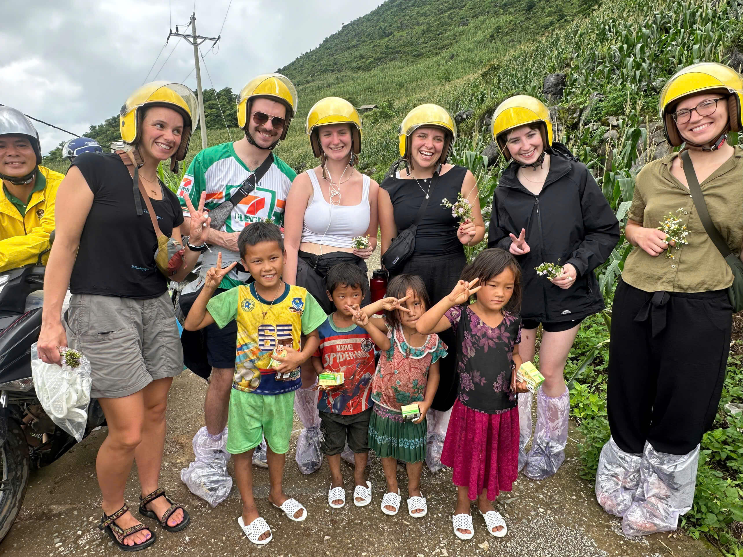 A small group of tourists wearing yellow motorbike helmets smiling and posing alongside local ethnic children on a quiet mountain road in Ha Giang