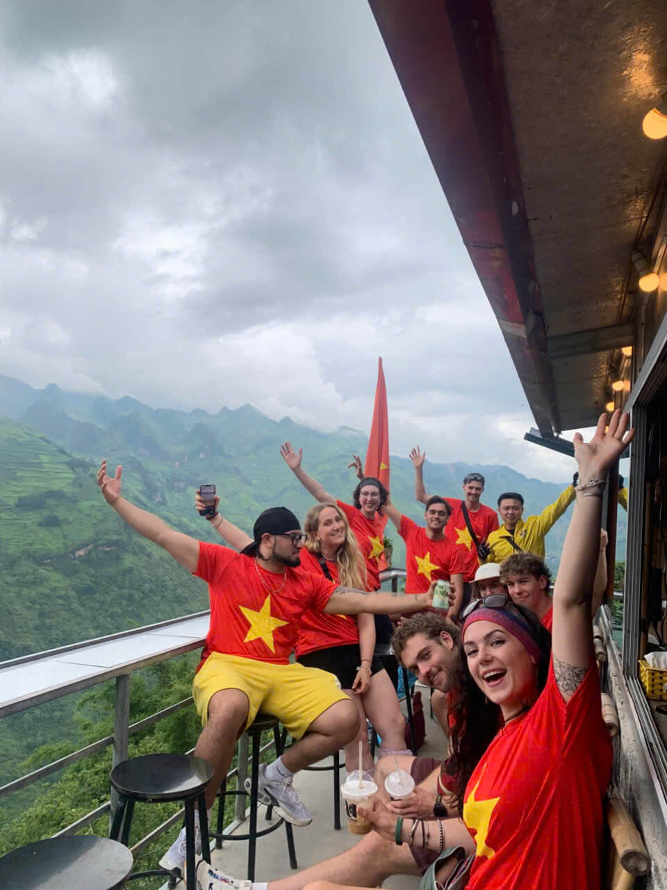 A tight-knit group of tourists wearing matching red Vietnam t-shirts cheering and celebrating together at a cliffside cafe with spectacular mountain views.