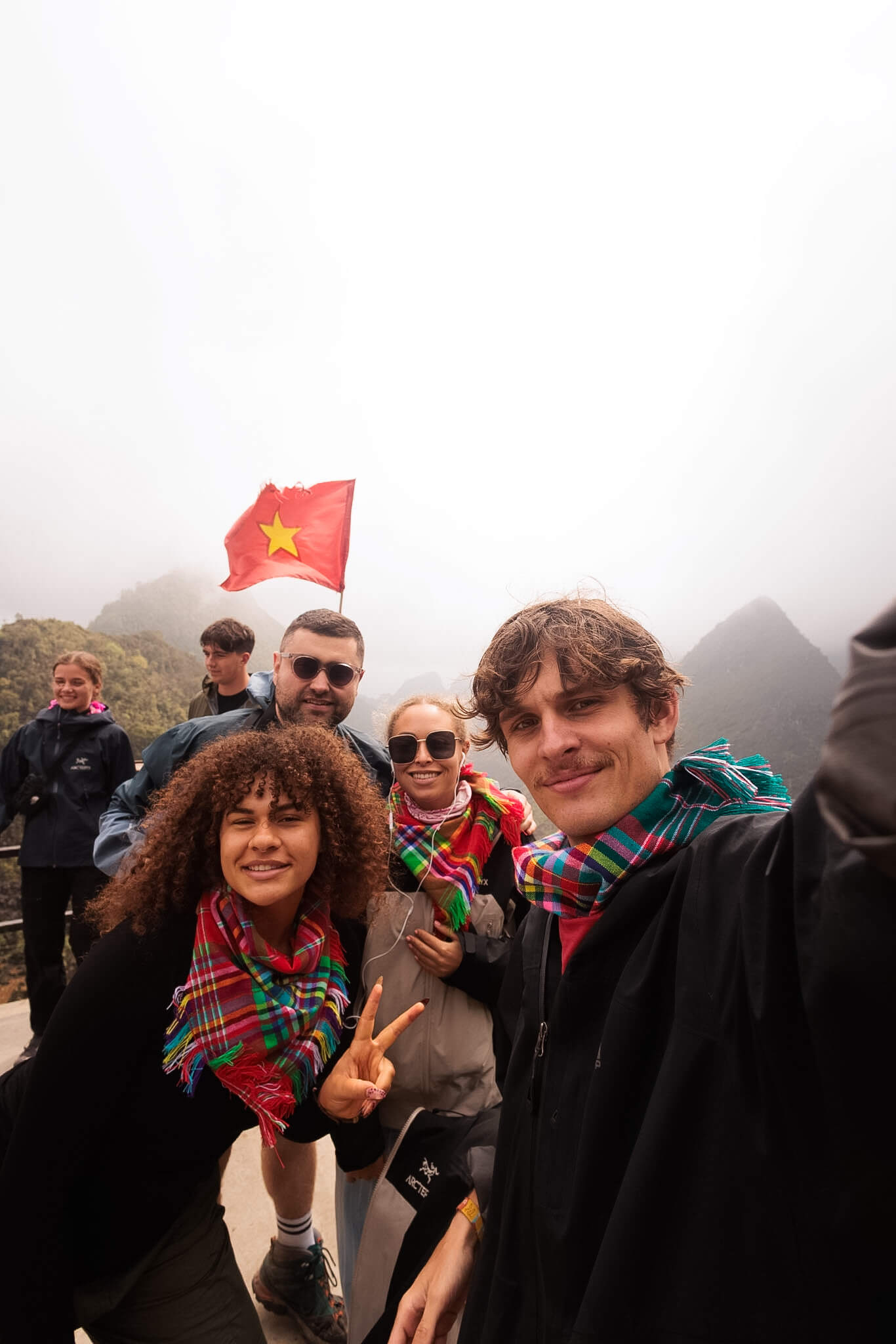 A group of cheerful young travelers wearing warm clothes and colorful ethnic scarves taking a selfie at a foggy mountain viewpoint in Ha Giang.