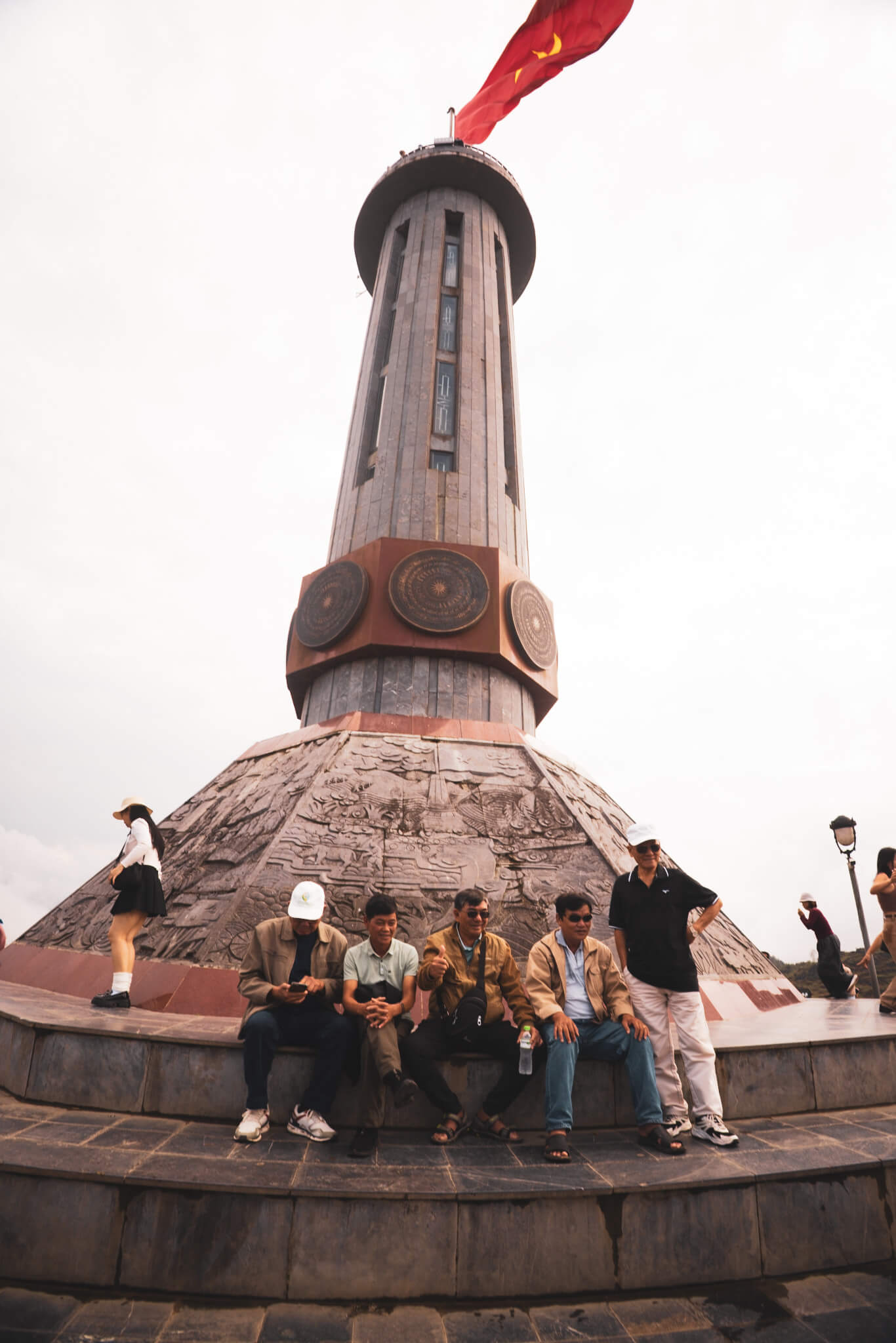 A group of visitors sitting at the base of the towering Lung Cu Flag Tower monument under a cloudy sky in Ha Giang, Vietnam.
