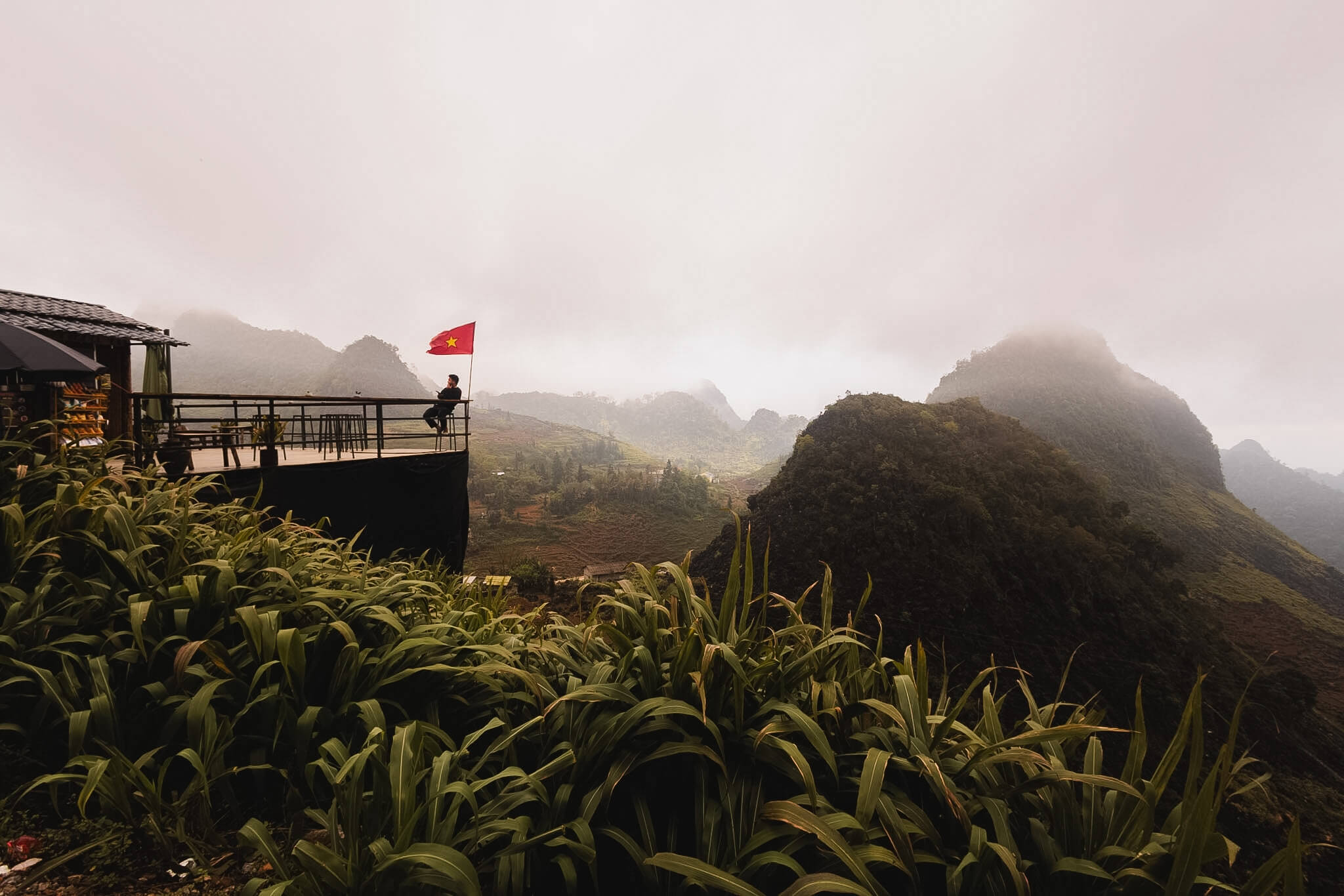 A cliffside observation deck with a red Vietnamese flag overlooking misty, fog-covered limestone karst mountains on the Ha Giang Loop.