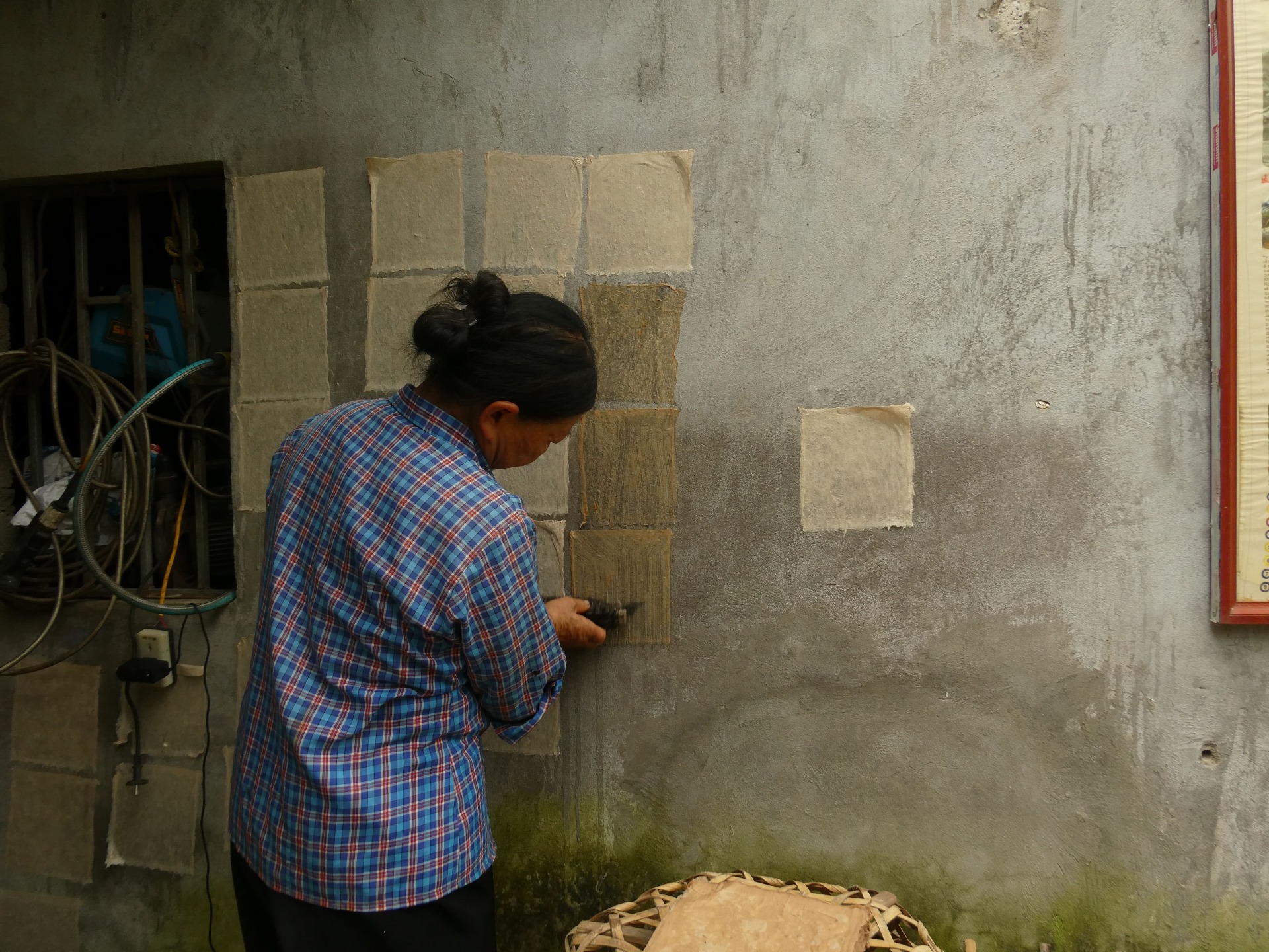 A local woman wearing a plaid shirt is carefully sticking individual squares of wet handmade paper onto a cement wall to dry.