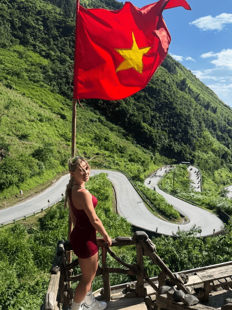 A female tourist in a red outfit standing on a wooden observation deck beneath a waving Vietnamese flag, looking out over a steep, winding mountain road featuring multiple hairpin turns and distant motorbikes.