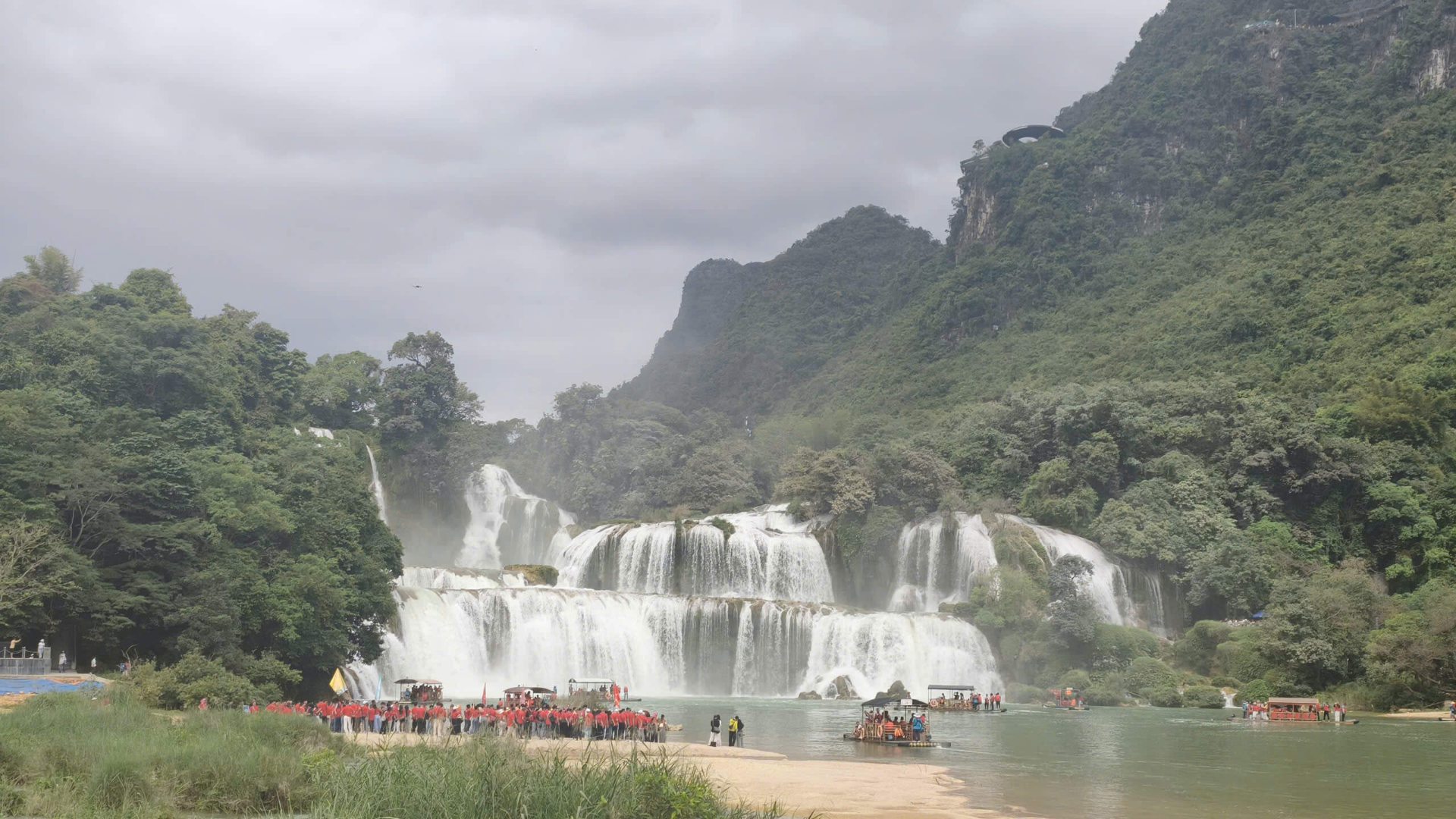 A bustling scene at Ban Gioc Waterfall with many tourists in red outfits gathering on the sandy riverbank on the left, while several tour boats and bamboo rafts navigate the green river waters under an overcast sky surrounded by lush, misty mountain cliffs in Cao Bang