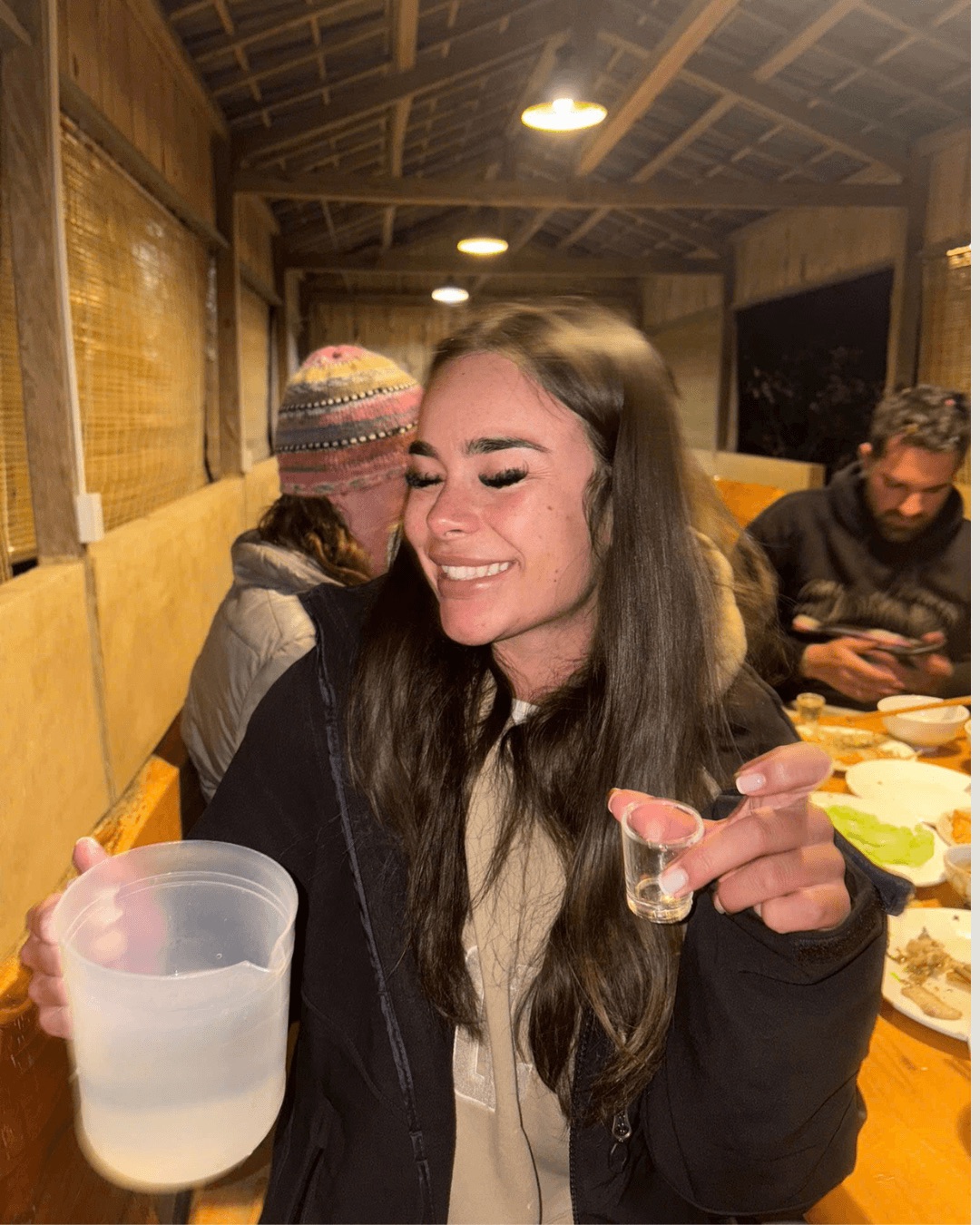 A happy female tourist with her eyes closed in laughter holding a small shot glass of happy water and a large plastic pitcher of rice wine during a communal dinner at a rustic homestay.