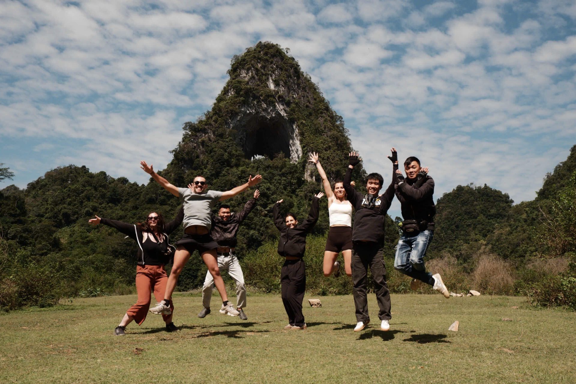 A group of six young people, including international tourists and a local guide, excitedly jumping into the air in a wide grassy meadow in front of Angel Eye Mountain in Cao Bang.