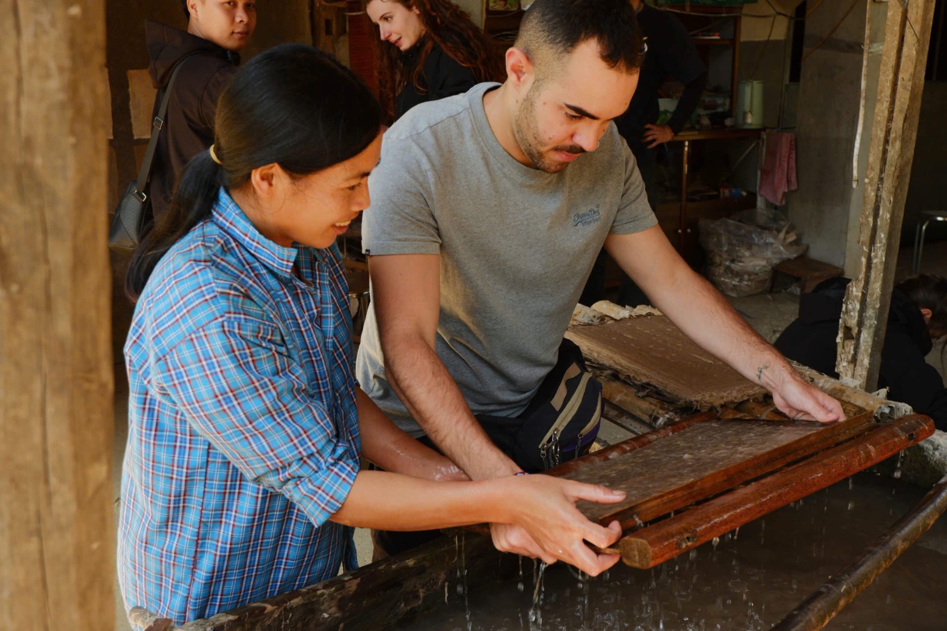 A local Nung woman in a plaid shirt guides a male tourist on how to lift a wooden frame to scoop natural fiber pulp from a vat during traditional paper-making in a Cao Bang craft village.