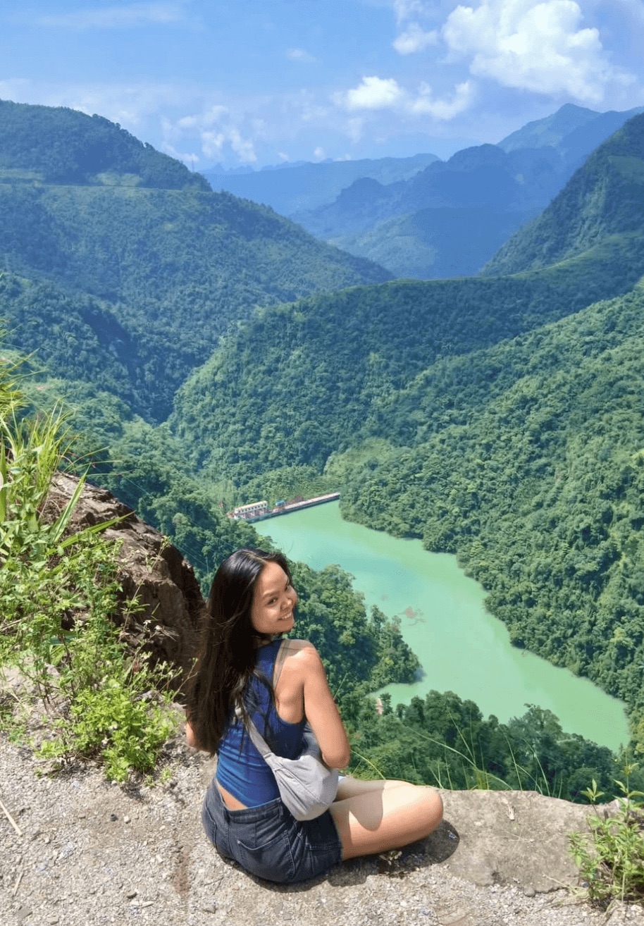 A female tourist wearing a small grey crossbody bag sitting on a rocky cliff overlooking the emerald green Nho Que River and surrounding mountains.