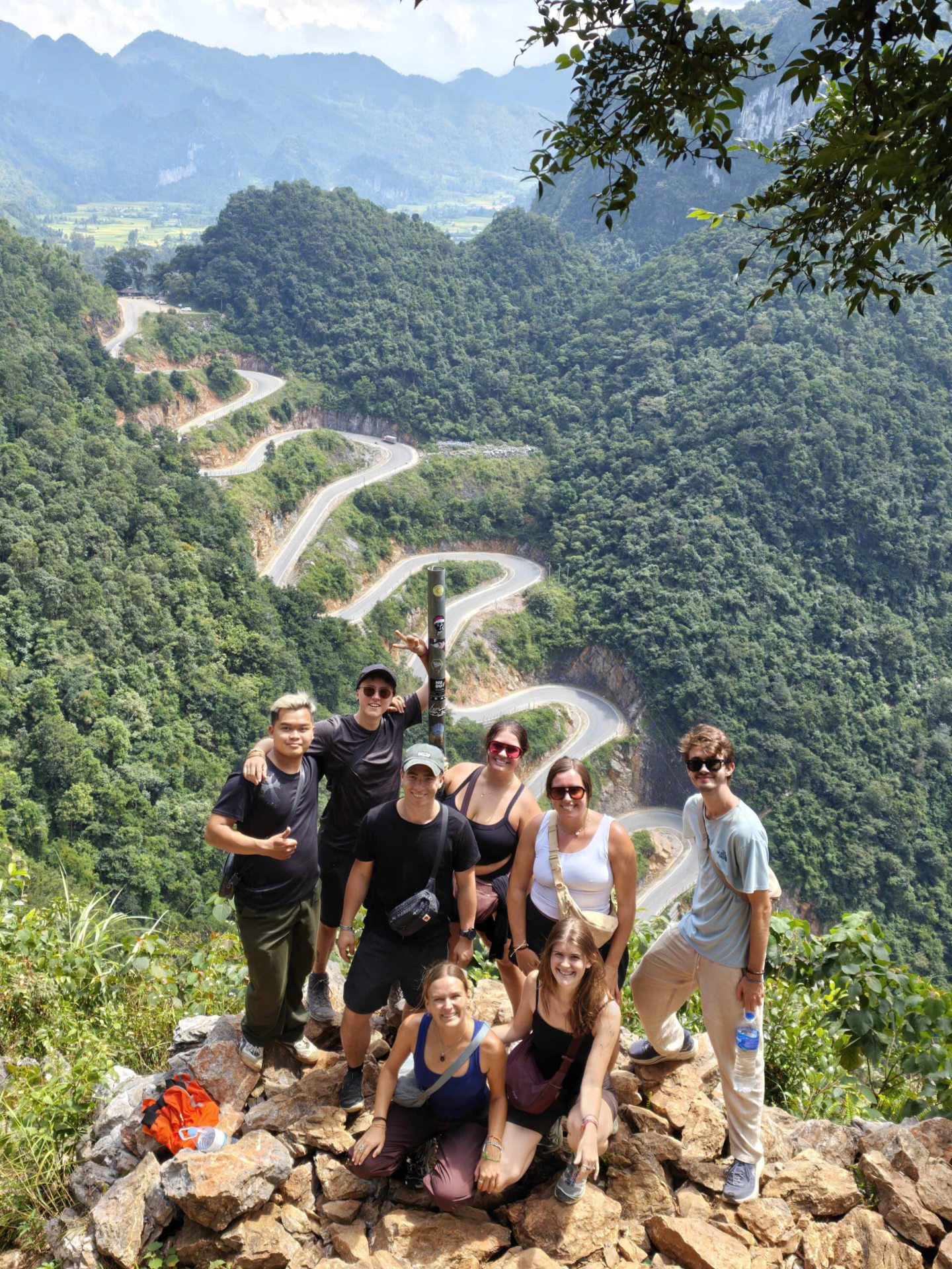 A large group of international tourists standing on a high rocky outcrop taking a group photo, with the spectacular 14-tier Me Pia mountain pass winding through the deep green forest in the background.