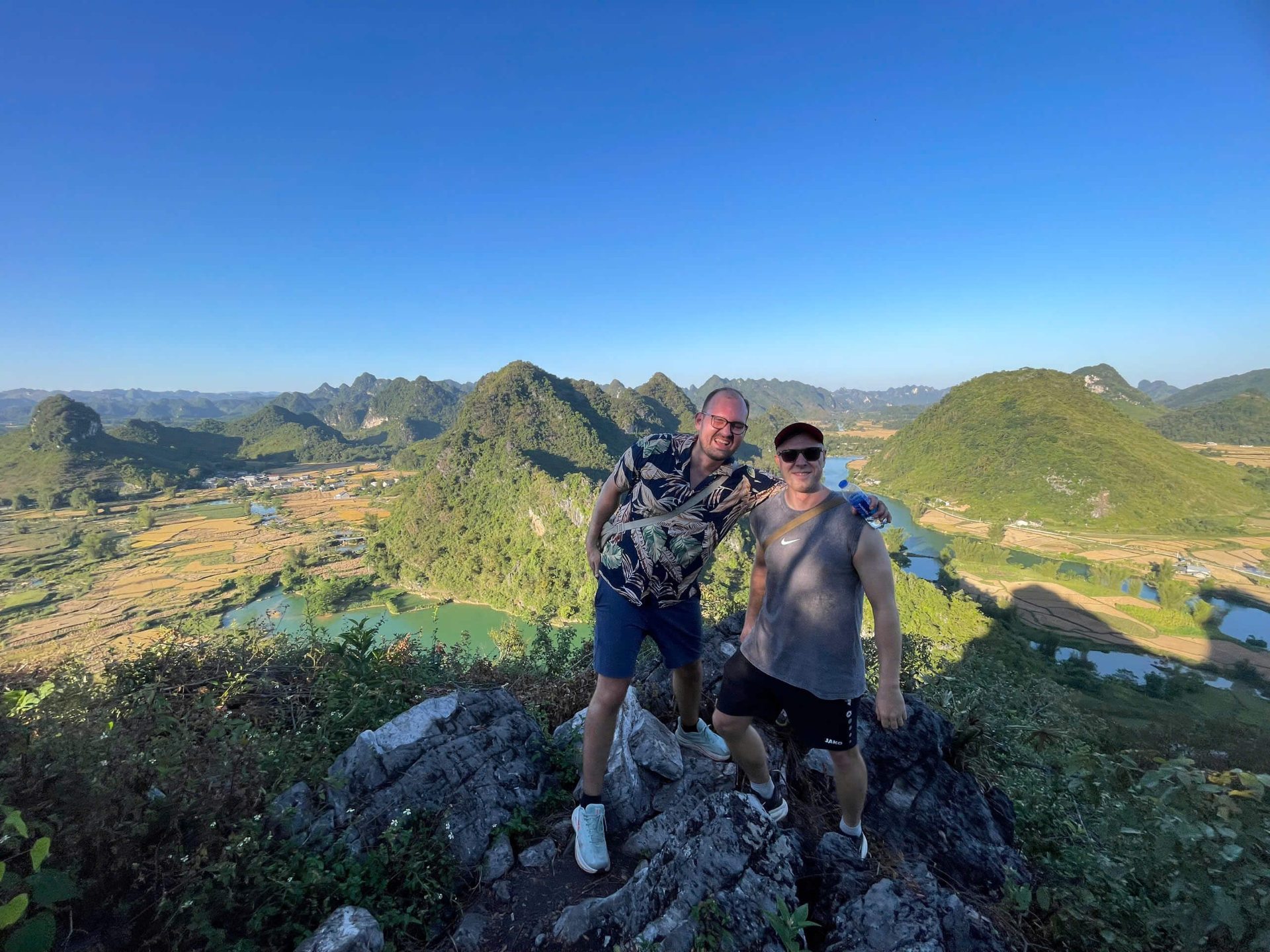 Two male tourists smiling and posing on a high rocky viewpoint overlooking the spectacular Phong Nam Valley, featuring a winding emerald river and golden rice paddies surrounded by lush karst mountains in Cao Bang.