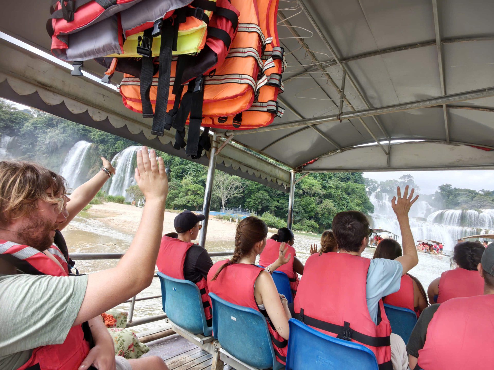 A group of tourists wearing orange and red life jackets seated on a covered tour boat, raising their hands and waving as they approach the roaring Ban Gioc Waterfall in Cao Bang