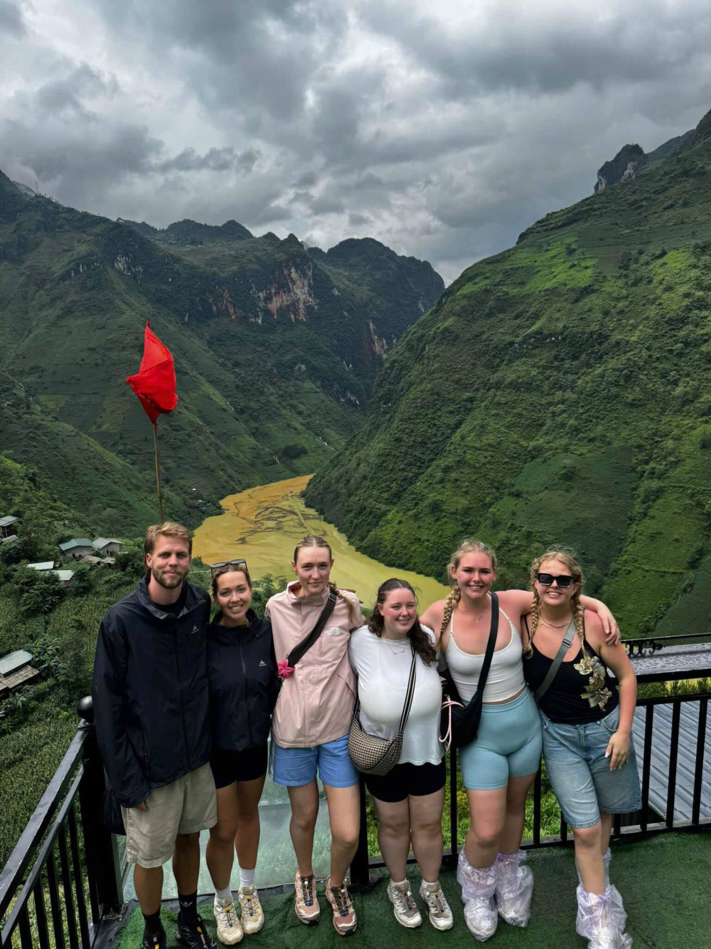 A happy group of international tourists and local easy rider guides wearing yellow shirts posing for a group photo on a wooden deck overlooking a vast mountain valley.